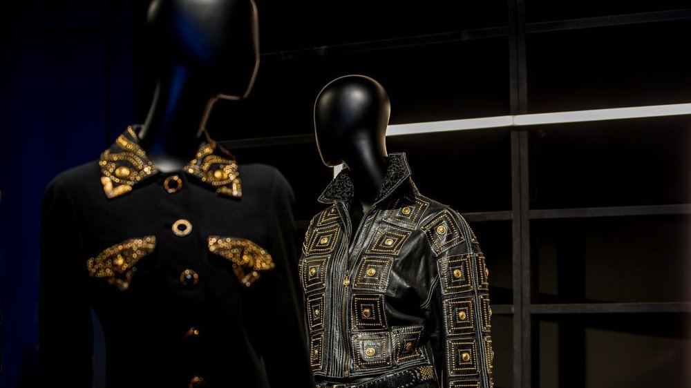 Dwa czarne manekiny, pierwszy prezentuje czarną koszulę ze złotymi kieszonkami oraz kołnierzem, drugi skórzaną, czarną kurtkę nabijaną złocistymi guzami i wyszywaną w złociste wzory.