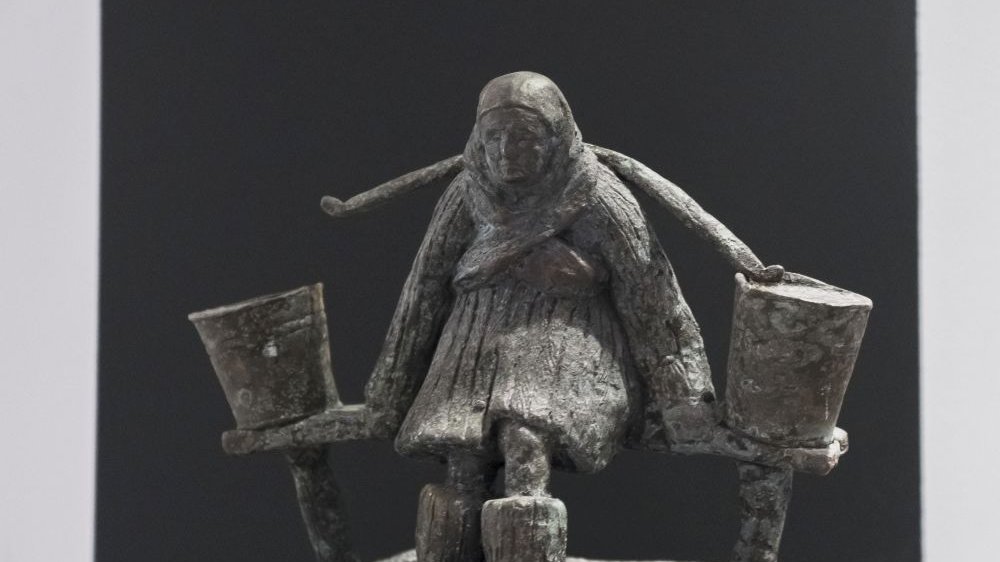 Rzeźba starej kobiety siedzącej na ławce, z nosidłem z dwoma wiadrami niesionym na plecach.