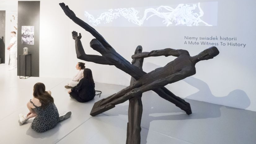 Widok ekspozycji - na środku kadru duża rzeźba w kolorze czarnym, obok niej, na podłodze, siedzą trzy kobiety, w tle ekran video.