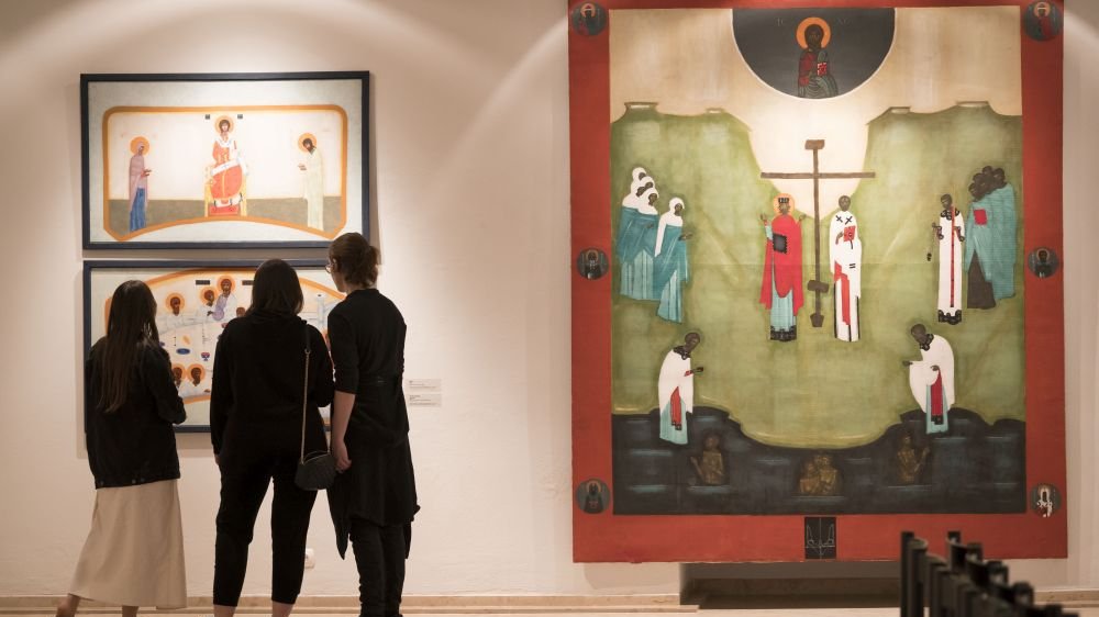 Trzy osoby ubrane w ciemne kolory oglądają obrazy na wystawie. Stoją przy mniejszym, białym obrazie w czarnych ramach.
