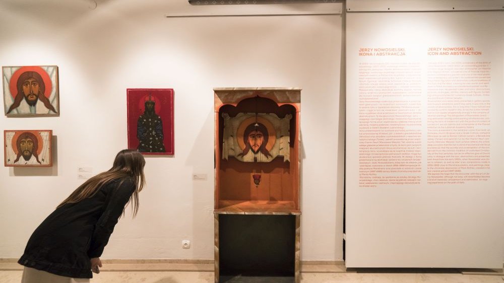 Zwiedzająca wystawę pochyla się obok jednej z ikon, umieszczonej w głębi drewnianego regału. Obok wiszą inne, małe ikony oraz opis wystawy.