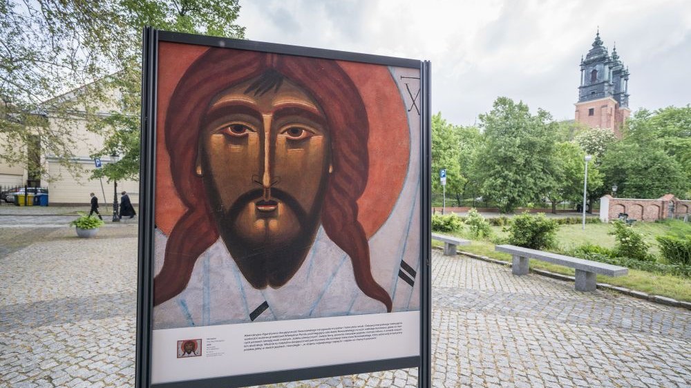 Potykacz informujący o wystawie, z namalowany wizerunkiem Jezusa. Stoi na Śródce, przed wejściem do muzeum, jest otoczony zielenią.
