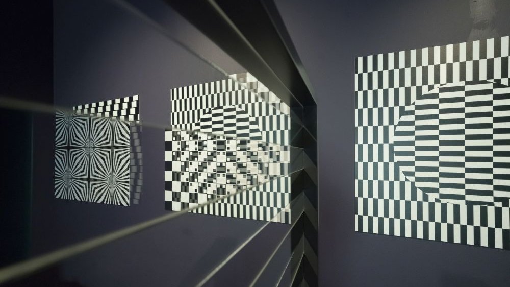 Trzy obrazy z czarno-białymi iluzjami optycznymi wiszą na ciemnej ścianie.