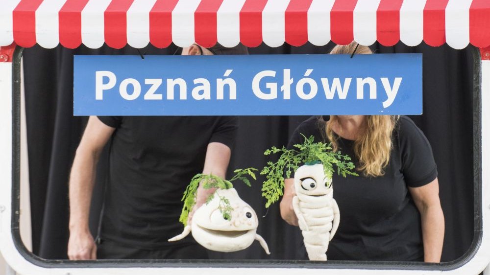 Aktorzy w czarnych koszulkach mają twarze zasłonięte napisem "Stacja: Poznań Główny". Najważniejsze na zdjęciu są lalki-warzywa - uśmiechnięty seler oraz pietruszka.