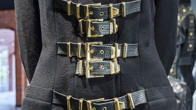 Czarny strój z dużą ilością skórzanych pasków ze złotymi klamrami prezentowany na manekinie.