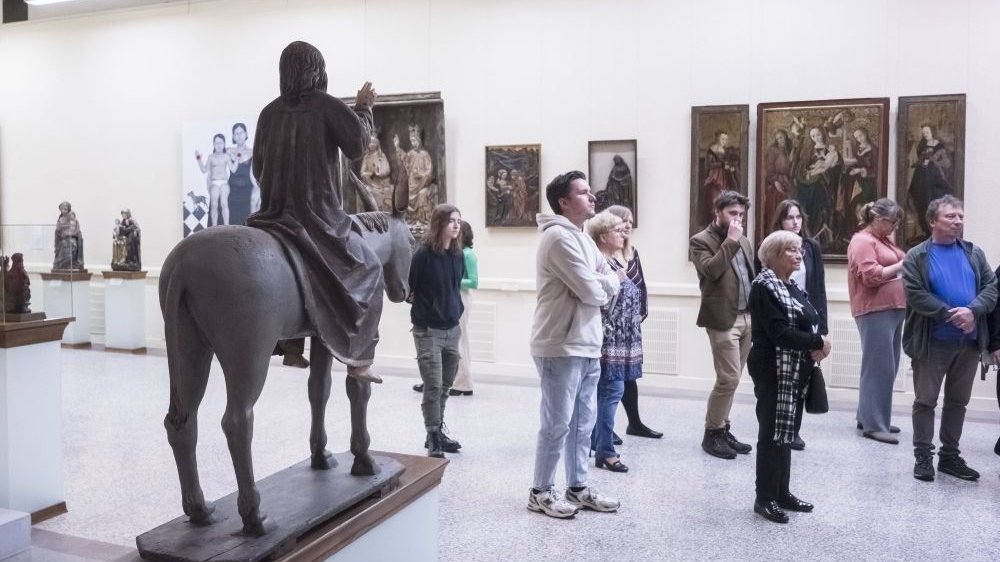 Grupa ludzi słucha przewodnika, obok nich stoi rzeźba Chrystusa na koniu. Za ludźmi wiszą obrazy z wystawy.