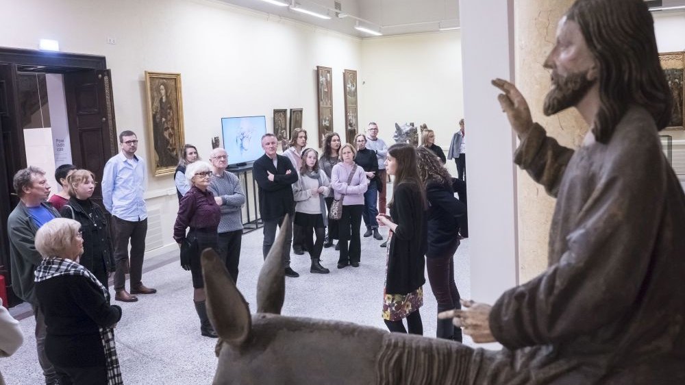 Widzowie wystawy przyglądają się wysokiej rzeźbie Chrystusa na koniu. Przewodniczki i kuratorki wystawy opowiadają.
