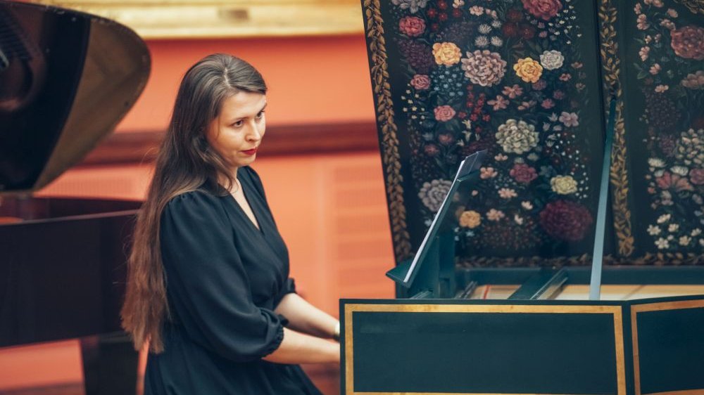 Kobieta w długich włosach zasiada przy pianinie z ozdobną klapą.