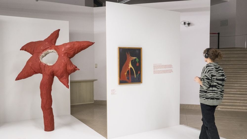 Dziewczyna ze słuchawkami na uszach zwiedza wystawę. Po lewej czerwona rzeżba przypominająca gwiazdę z dziurą w środku stojąca na jednej nodze.