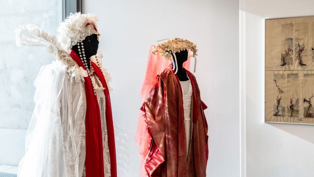 Dwa kostiumy teatralne na manekinach. Powłóczyste szaty z dodatkami pereł, sztucznych kwiatów i piór.