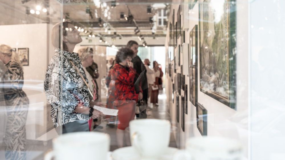 Ludzie zwiedzają wystawę, zdjęcie zrobiono zza szyby gabloty, w której stoją białe porcelanowe filiżanki.
