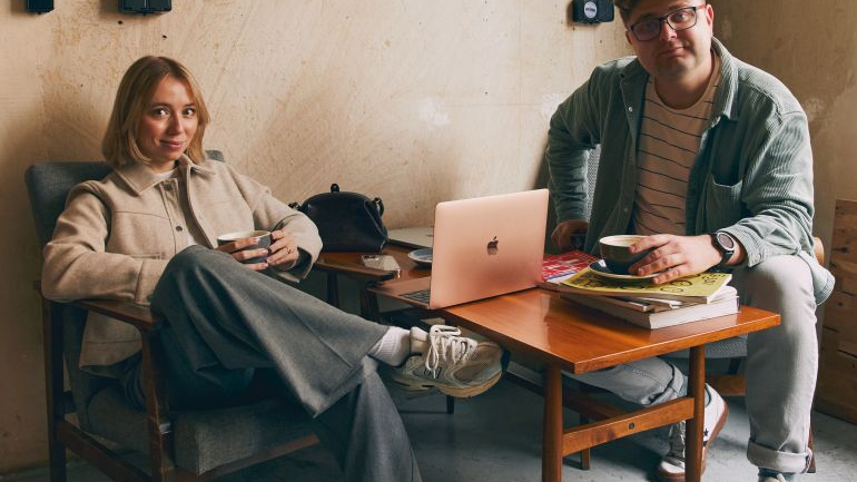 Para siedzi w kawiarni przy stoliku, na którym leży otwarty laptop oraz kilka książek. Na ścianie za nimi wisi kolorowy obraz.