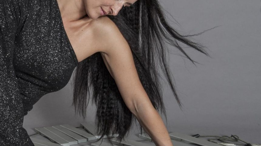 Długowłosa artystka w błyszczącej, czarnej sukni pochyla się ku podłodze i dotyka leżącego na niej instrumentu.