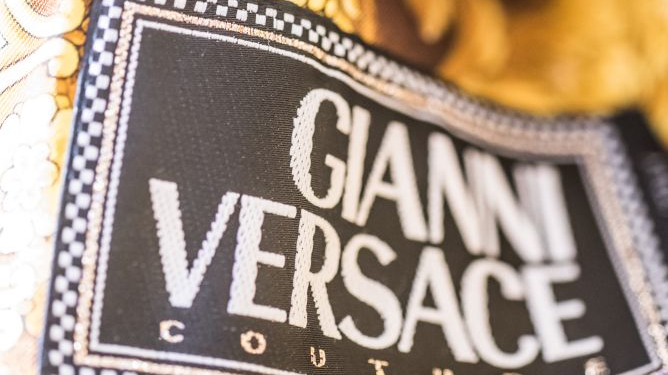 Zbliżenie na metkę "Gianni Versace Couture" wszytą w złotą, wzorzystą tkaninę.