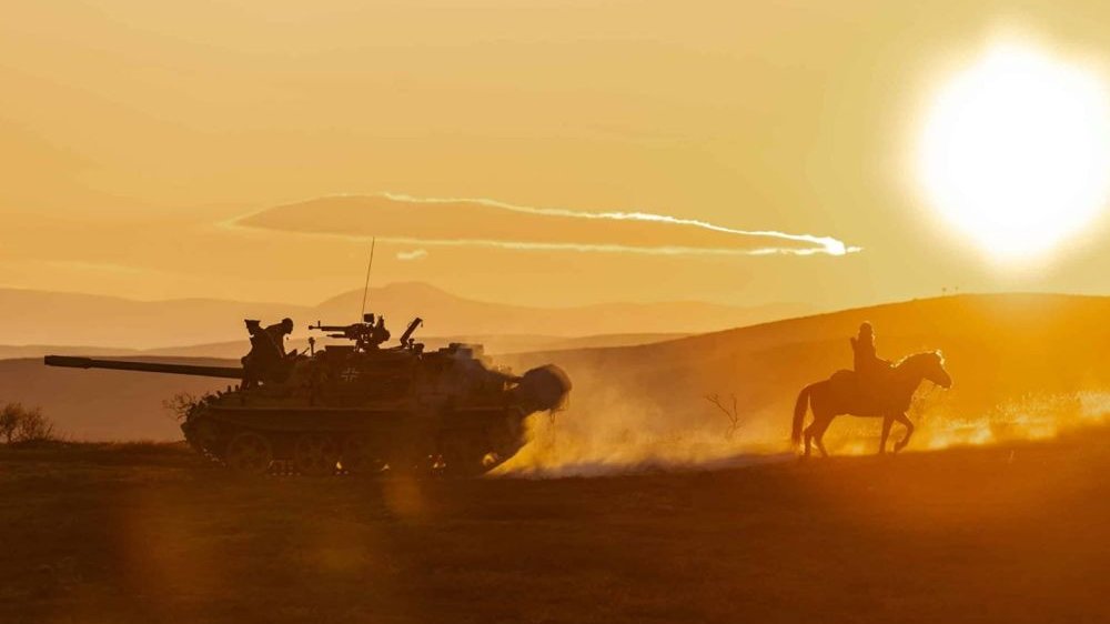 Jeździec na koniu, za nim czołg. Jadą przez pustynię w czasie zachodu słońca.