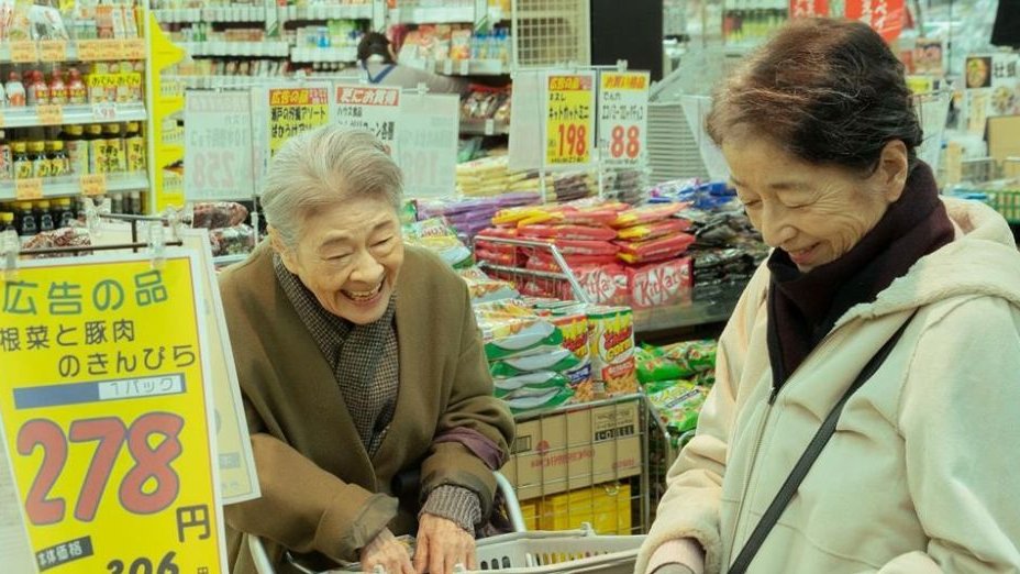 Dwie starsze kobiety w sklepie spożywczym. Obie się śmieją. Z tyłu kolorowe produkty na półkach.