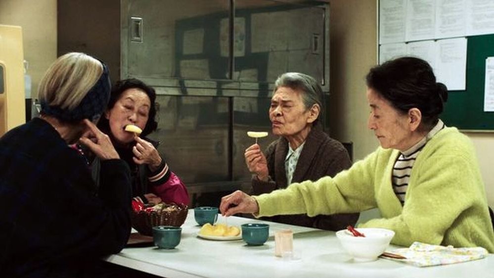 Grupa starszych ludzi siedzi przy stole, jedzą coś wykałaczkami. Z tyłu za nimi metalowa szafka i zielona tablica z ogłoszeniami.