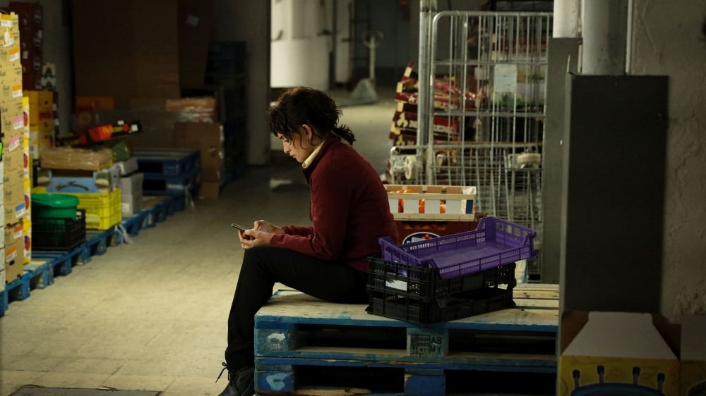 Kobieta w bordowej bluzie siedzi na drewnianej palecie na zapleczu sklepu. Ma opuszczoną głowę, ogląda coś na ekranie swojego smartfona.