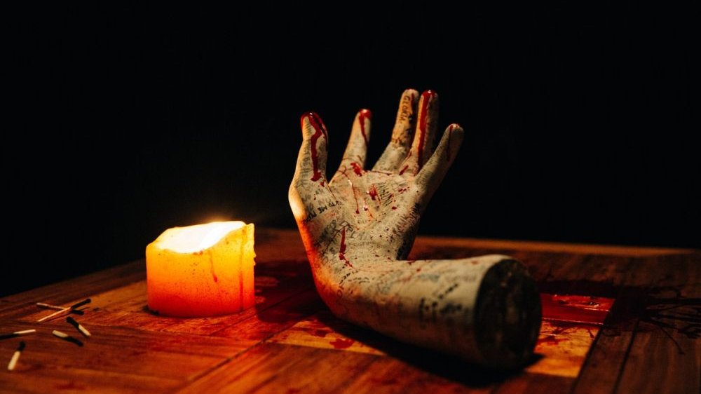 Zakrwawiona i wytatuowana zabalsamowana ludzka ręka leży na stole w nienaturalnej pozie. Obok niej płonąca świeca i zużyte zapałki.