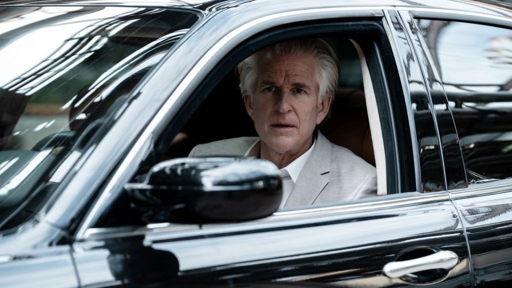 Z okna samochodu, miejsca kierowcy, wygląda siwy, starszy mężczyzna w jasnoszarym garniturze.