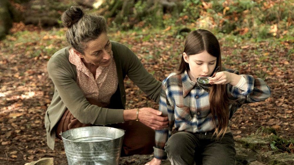 Dziewczynka w kraciastej koszuli pije wodę z chochli, za ramię trzyma ją starsza kobieta. W tle jesienny krajobraz.