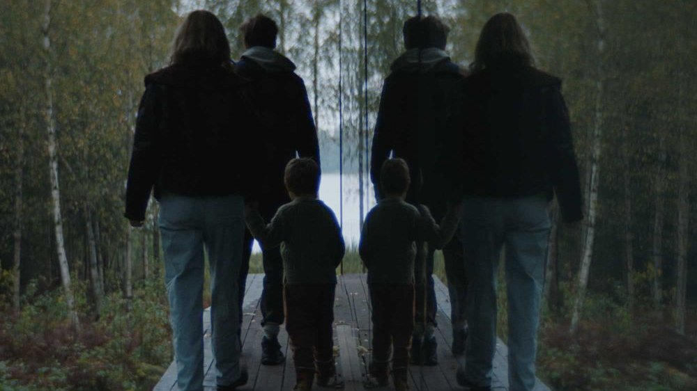 Rodzice wraz z synkiem idą drewnianą ścieżką, z boku odbija ich lustro, przez co wydają się "podwojeni", "skopiowani".