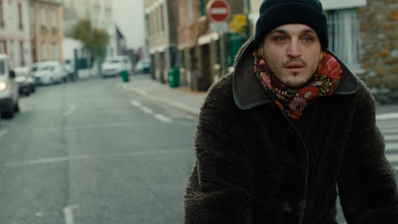 Bohater ubrany w futrzaną kurtkę, szalik w kwiaty i czarną czapkę idzie pustą ulicą w ciągu dnia.