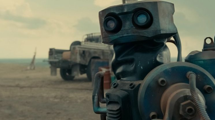 Metalowy robot z dwoma szkiełkami w miejscu oczu, za nim pustkowie i samochód.