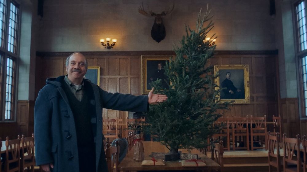 Mężczyzna z wąsem dumnie prezentuje choinkę. Drzewko stoi na stole, na ścianach obrazy i głowa jelenia.