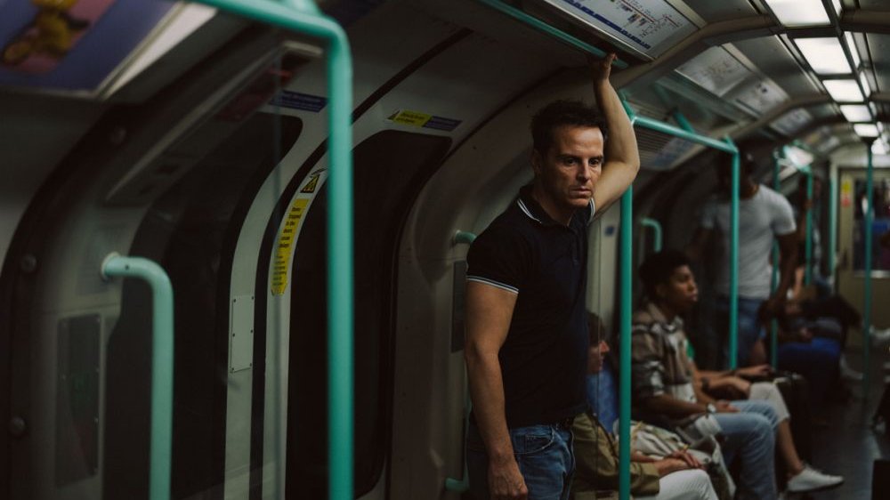 Mężczyzna podróżuje metrem, stoi trzymając się jedną ręką drążka w górze. Jest zamyślony.