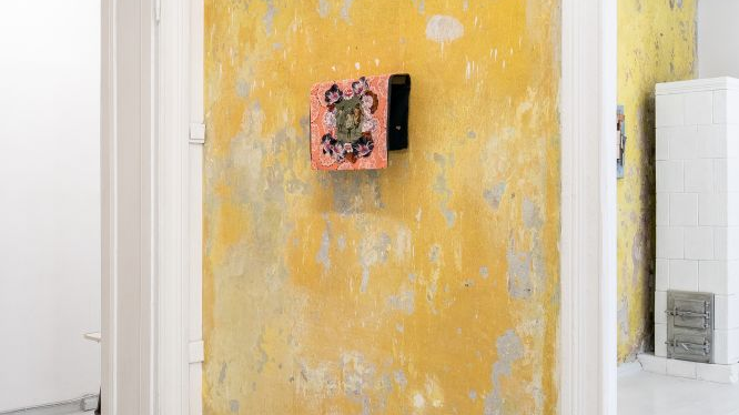 Praca z kolorową makatką powieszona na żółtej, odrapanej ścianie galerii. Z tyłu, w drugim pomieszczeniu, biały piec kaflowy.