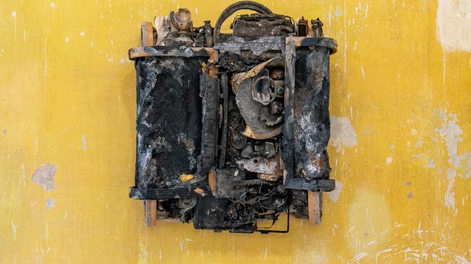 Złożony z różnych elementów obiekt powieszony na żółtej ścianie. Sprawia wrażenie spalonego sprzętu elektronicznego lub części jakiejś maszyny.