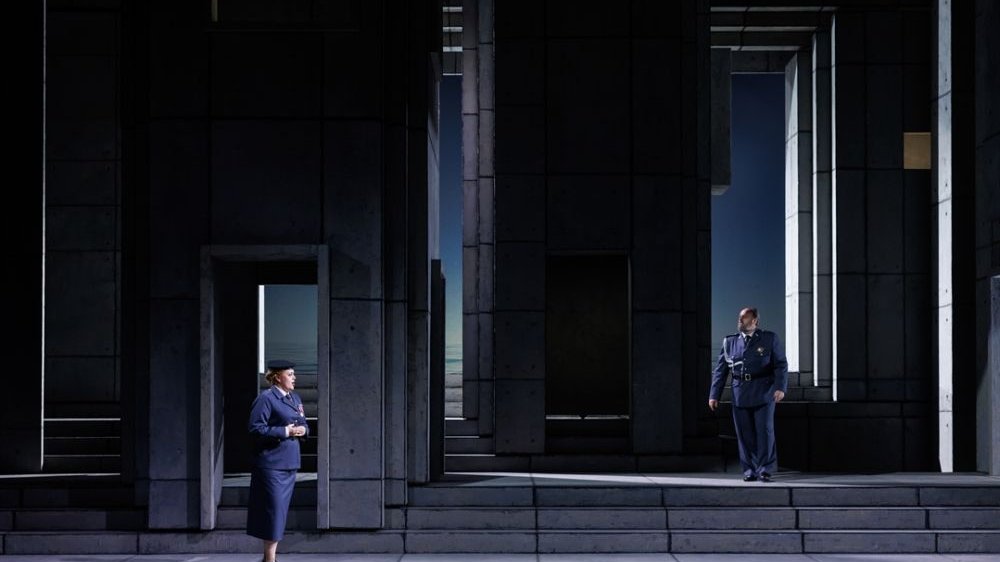 Kobieta i mężczyzna w mundurach wojskowych stoją na scenie, w surowej, szarej scenografii.