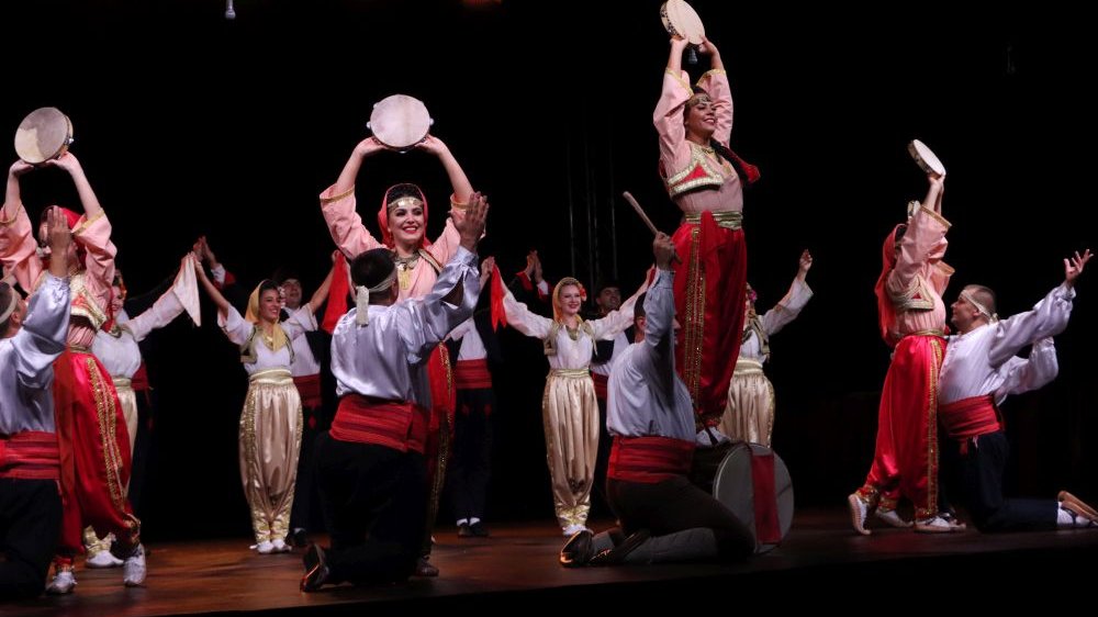 Tancerki w szerokich spodniach w kolorach czerwonym i złotym stoją w kilku rzędach. Tancerki w pierwszym rzędzie grają na tamburynach i sa podtrzymywane przez tancerzy.