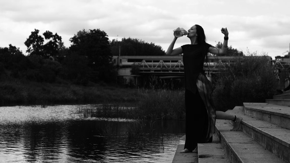 Czarno-biała fotografia, kobieta w czarnym stroju stojąc nad brzegiem rzeki dmucha w muszlę. W tle widać stalowy most.
