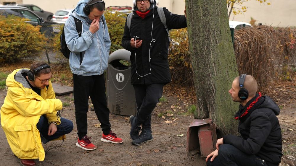 Grupa ludzi w słuchawkacj na uszach stoi lub kuca przy metalowej skrzynce tuż obok pnia drzewa.