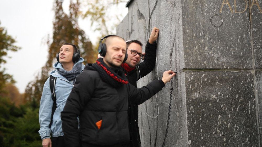 Trzech mężczyzn w słuchawkach stoi obok pomnika Adama Mickiewicza. Dwóch z nich przykłada do niego kable.