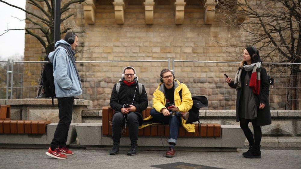 Czworo młodych ludzi stoi lub siedzi przed CK Zamek w Poznaniu. Słuchają nagrania audio na swoich telefonach.