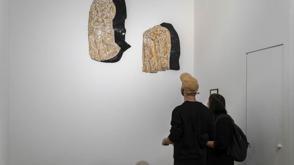 Para zwiedzajaca galerię przygląda się kompozyzcji dwóch dzieł, przypominających kamienie, wywieszonych obok siebie na białej ścianie.