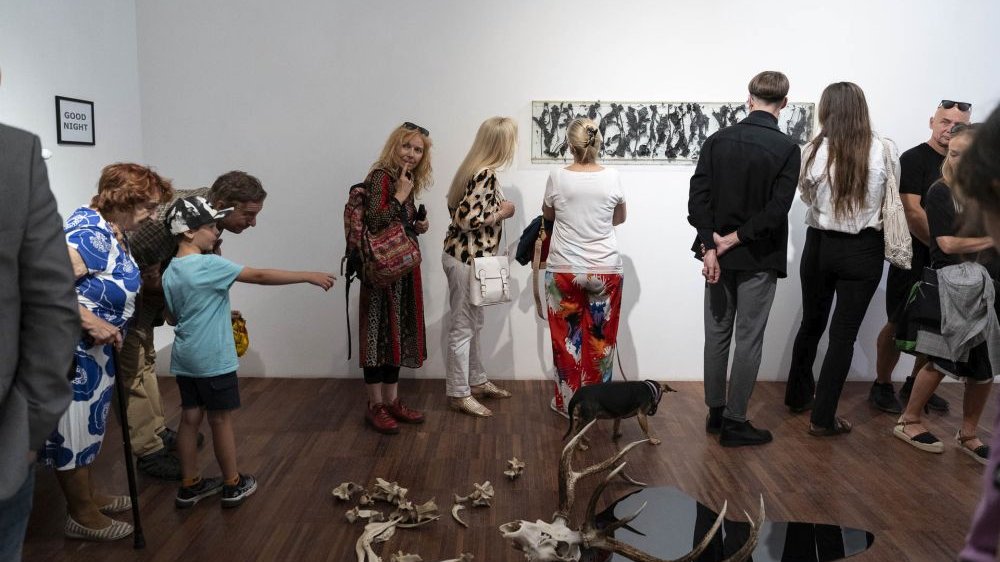 Grupa ludzi zwiedza wystawę podczas wernisażu, rozglądając się po galerii. Na podłodze leży kompozycja z kości i czaszki z porożem, na ścianie wisi czarno-białe dzieło sztuki.