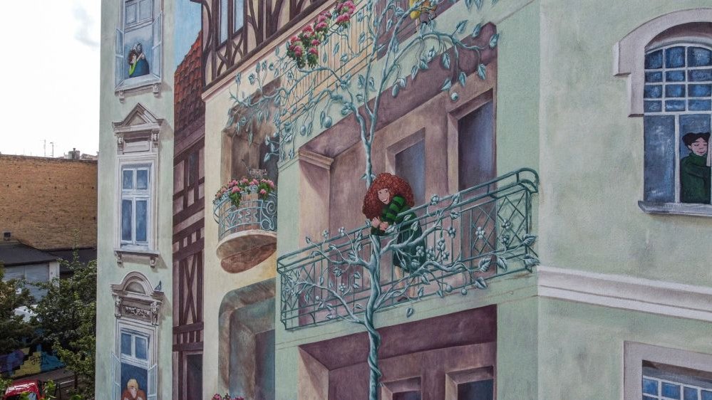 Fragment muralu, na którym widać wychylającą się przez balkon i szeroko uśmiechniętą rudą dziewczynę o bujnych włosach, w zielonej sukience w czarne paski.