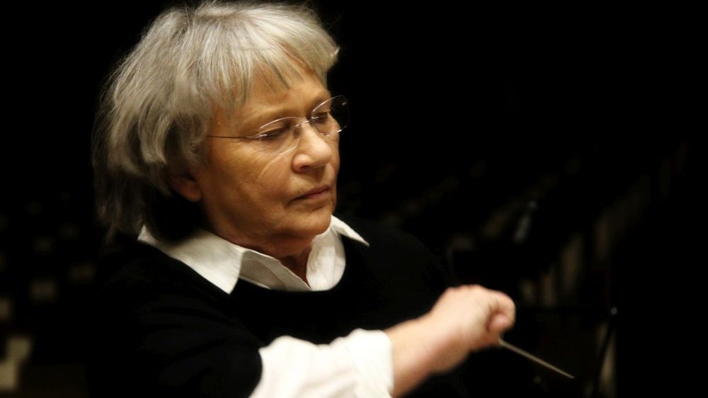 Agnieszka Duczmal dyryguje orkiestrą, ma zamknięte oczy. Za nią ciemność.