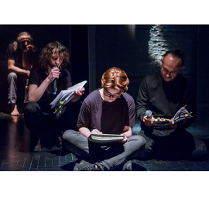 Kilku mężczyzn siedzi na scenie, każdy z nich trzyma przed sobą otwartą książkę. Jeden z mężczyzn patrząc na otwartą książkę czyta do mikrofonu.