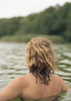Kobieta w wodzie, widać plecy i długie włosy. Z przodu las.