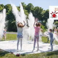 Fotografia przedstawia dzieci rzucające białym proszkiem ponad białym materiałem rozłożonym na trawie. Dodatkowo na fotografię nałożono logo 21. edycji Letniego Pogotowia Sztuki.