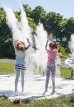 Fotografia przedstawia dzieci rzucające białym proszkiem ponad białym materiałem rozłożonym na trawie. Dodatkowo na fotografię nałożono logo 21. edycji Letniego Pogotowia Sztuki.