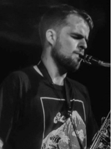 Czarno biała fotografia przedstawia profil młodego mężczyzny grającego na saksofonie