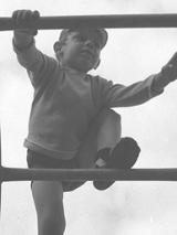 Czarno-biało fotografia przedstawiająca chłopca, który wspina się po drabinie. Na nogach ma wysokie podkolanówki. Nad nim widać zachmurzone niebo.