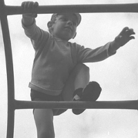 Czarno-biało fotografia przedstawiająca chłopca, który wspina się po drabinie. Na nogach ma wysokie podkolanówki. Nad nim widać zachmurzone niebo.