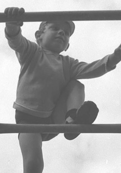 Czarno-biało fotografia przedstawiająca chłopca, który wspina się po drabinie. Na nogach ma wysokie podkolanówki. Nad nim widać zachmurzone niebo.